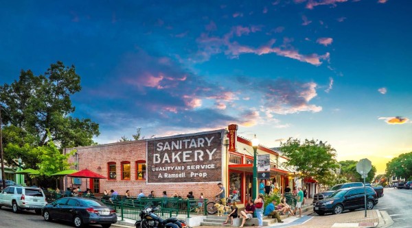 A vibrant streetscape at dusk showing a low brick building with a large painted sign that reads “SANITARY BAKERY – QUALITY AND SERVICE A. Ramsell, Prop.” Red-trimmed windows and a small “PIZZERIA” blade sign flank its corner entrance. In front, people sit at outdoor tables under red and green umbrellas, while others chat on a raised patio platform. A black motorcycle and several cars line the street beside a raised curb and crosswalk. Lush trees frame the scene under a deepening blue sky streaked with pink-tinged clouds.