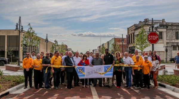 A large group of people gathered in downtown Denison, Texas, for a ribbon-cutting ceremony. They are holding a banner that reads 'Congratulations!' while standing on a newly renovated brick-paved street. The group includes community leaders, local officials, and residents, with a mix of yellow shirts, business attire, and casual wear. The setting is framed by restored historic buildings, trees, and a stop sign marking Main Street and Rusk Avenue.