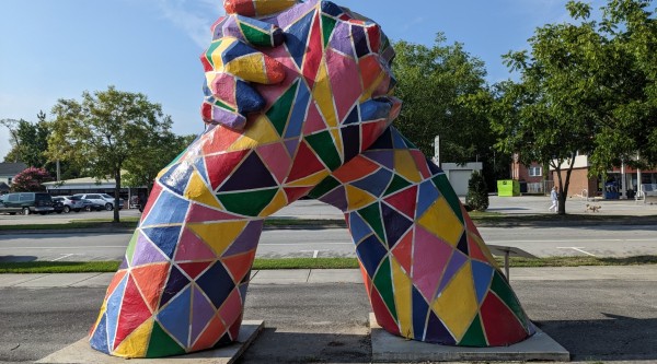 A large public sculpture of two oversized legs rising from square concrete bases and meeting at the top in a firm handshake. The entire piece is painted in a vibrant mosaic of geometric shapes—triangles and quadrilaterals in red, yellow, green, blue, pink, and purple—outlined in white. It stands on a roadside sidewalk with a parking lot, trees, and low buildings visible in the background.