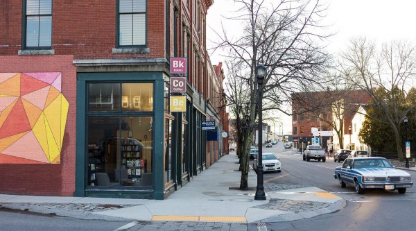 A quiet downtown corner at dusk shows a red-brick building with large display windows revealing bookshelves inside. To the left, a colorful geometric mural in shades of pink, yellow, and orange fills the lower brick wall. Vertical signs above the doorway read “Bk Books,” “Co Coffee,” and “Be Beer.” A leafless street tree and vintage streetlamp stand on the wide concrete sidewalk, while a blue-and-white classic sedan and other cars drift past on the two-lane road flanked by more historic storefronts.