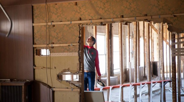 A person in a red-and-gray pullover stands inside a partially demolished interior, holding a white hard hat and looking up at exposed wooden studs and wall framing. The room has drywall removed from one side, a red ladder lying on the floor, and sunlight streaming through tall windows in the background.