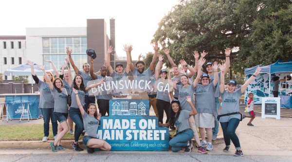 A group of about twenty people wearing matching “Made on Main Street” t-shirts stand and kneel behind a layered sign reading “Brunswick, GA” over “Made on Main Street – communities. Celebrating each other.” They’re outdoors on a street curb with event tents and a modern building behind them, and nearly everyone has arms raised in an enthusiastic gesture.