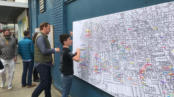 People in Covington, Kentucky add color-coded stickers to a large illustrated map mounted on a downtown building wall as part of a community engagement activity.