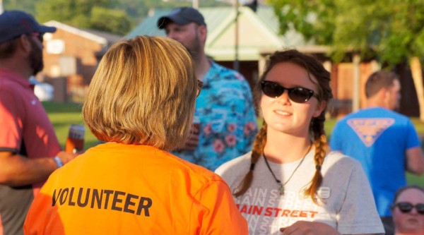 A person wearing a bright orange shirt with “VOLUNTEER” printed in bold on the back talks to someone facing the camera who is wearing sunglasses and a gray “Main Street” t-shirt; they stand under a canopy at an outdoor event with other attendees mingling in the background.
