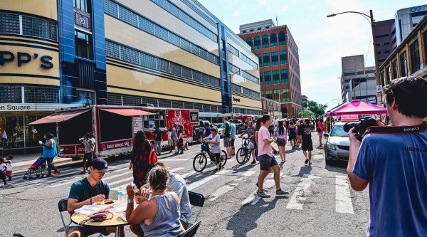 A sunny city street closed to traffic hosts a lively community event. In the foreground, three people sit at a small round table eating from take-out containers while another person at right films the scene with a DSLR camera. Behind them, attendees stand in line at a red “Award Winning BBQ” food truck, and pedestrians and bicyclists wander past art-deco–style and brick commercial buildings under a partly cloudy sky.