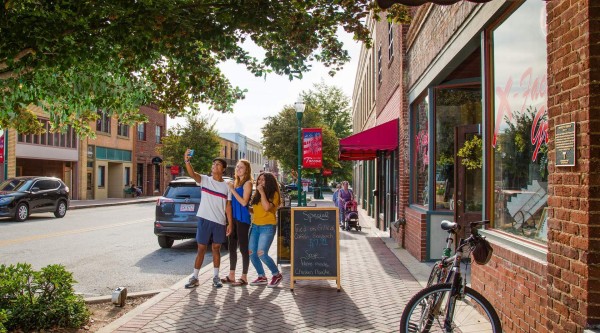 Three people stand on a brick-paved sidewalk beneath leafy tree branches, holding up a phone together to take a selfie. To their right, a sandwich-board sign advertises specials in front of a red-awning storefront, and a bicycle leans against the building. Parked cars line the street, and a parent pushes a stroller farther down the sidewalk under a banner–draped lamppost.