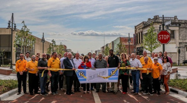 A large group of people stands across a freshly brick-paved street in front of new streetlights and storefronts. In the center, two individuals hold oversized ceremonial scissors poised over a yellow ribbon printed with “Congratulations!” as others grasp the ribbon at either end. Many in the crowd wear matching gold shirts, while others are dressed in business or casual attire. Young trees line the street, and a stop sign and “Main St” street sign are visible on the right.
