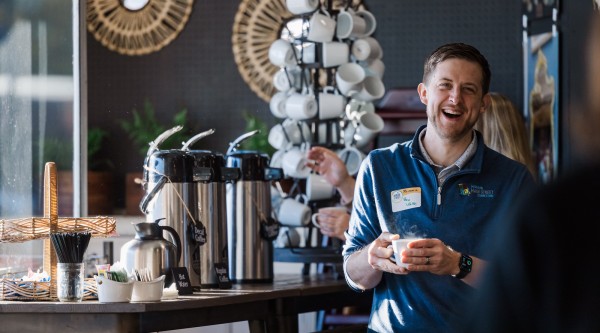A smiling man wearing a blue Missouri Main Street Connection pullover and a name badge stands near a coffee station, holding a steaming white mug. Behind him, a rotating mug rack and wicker baskets of stirrers and condiments sit atop the counter, while decorative woven wall mirrors and natural light fill the cozy café setting.