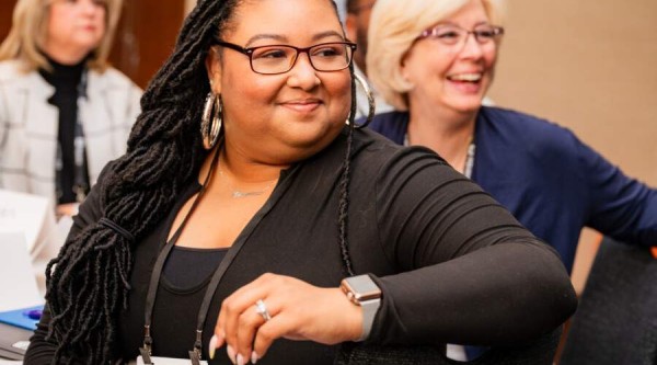 A conference attendee with long braided hair, wearing glasses, hoop earrings, and a black top, sits at a table smiling and looking to her left. She has a name badge around her neck and a smartwatch on her wrist. Behind her, another attendee with short blonde hair and glasses laughs, and a third person sits further back in a patterned jacket.