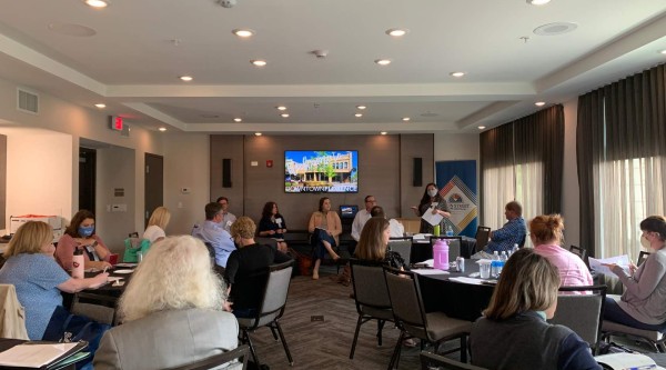 A small conference room set up with round tables where about a dozen participants sit facing the front. At the head of the room, five panelists are seated in front of a large wall‐mounted screen displaying “Downtown Florence,” and a masked presenter stands beside them holding notes. Attendees around the tables are reviewing papers, sipping water, and listening—some wearing masks—and laptops, notebooks, and beverage bottles are spread across the tables.