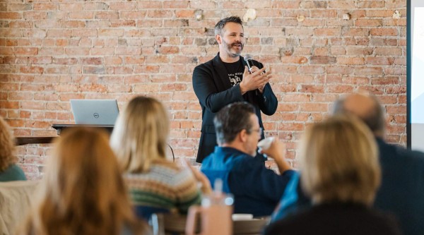 A man stands at the front of a room holding a microphone and gesturing as he speaks. He wears a black blazer over a dark t-shirt and stands in front of an exposed brick wall. In the foreground, several seated attendees—seen from behind—listen, one sipping from a cup. A laptop sits on a small table beside the speaker.