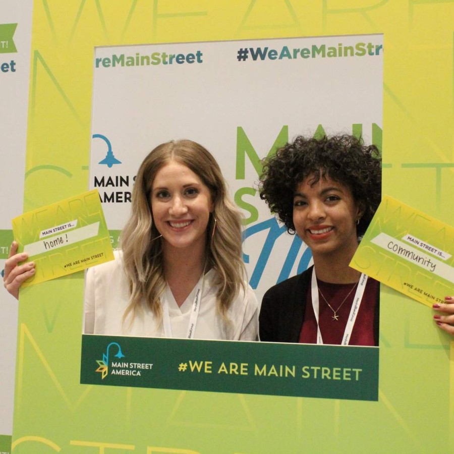 Two smiling women pose behind a large lime-green frame branded “Main Street America” and “#WeAreMainStreet.” Each holds a small card: the woman on the left’s reads “Main Street is… home!” and the woman on the right’s reads “Main Street is… community.” Behind them is a repeating backdrop celebrating 40 years of the Main Street movement.