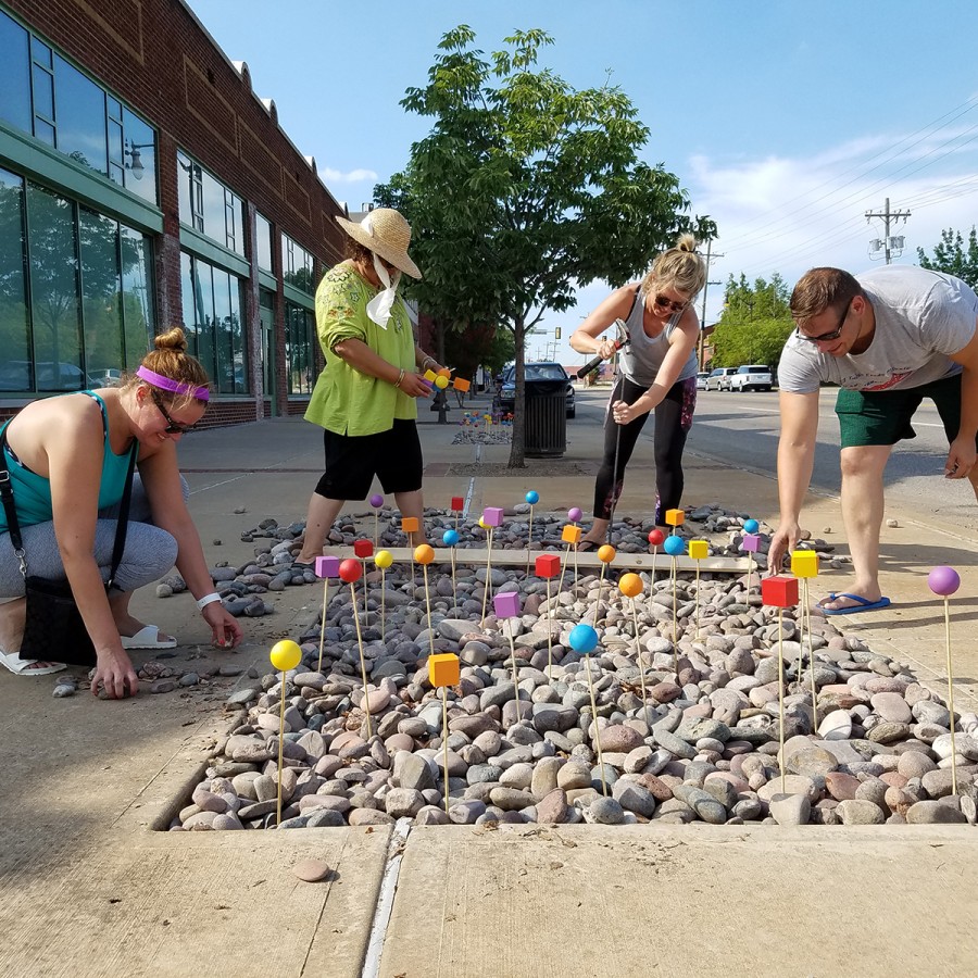 A group of four people works on a sidewalk art installation set within a recessed bed of river rocks. Dozens of slender wooden stakes topped with painted wooden shapes (spheres and cubes in bright yellow, orange, red, blue, and purple) rise from the stones. On the left, a crouching woman in a teal tank top and gray leggings picks up rocks; next to her, a standing woman in a wide-brimmed straw hat and green shirt holds two yellow-and-orange blocks. On the right, a woman in a gray tank top drives a stake into the stones with a mallet, while a man in a gray t-shirt and green shorts bends over to adjust a red cube on its stake. Behind them, a row of brick storefronts and the street with parked cars frame the scene under a clear blue sky.