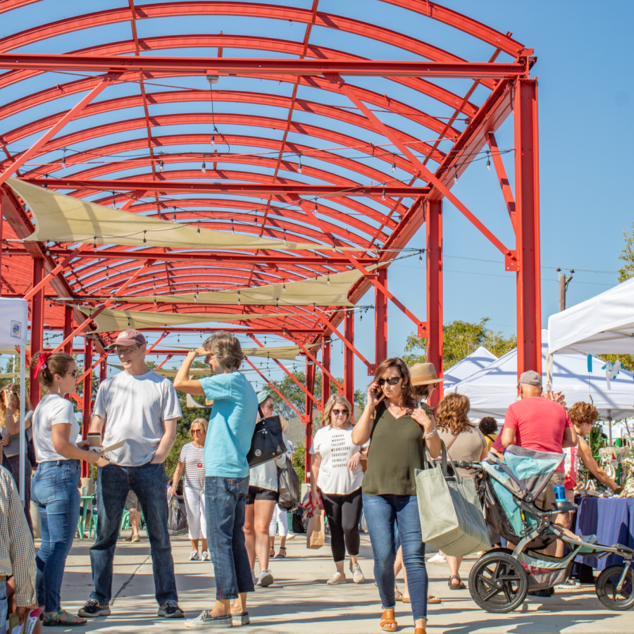 On a bright and sunny day, people shop at a farmers' market. A vibrant red archway spans the pavilion where vendors are set up under pop-up canopy tents.