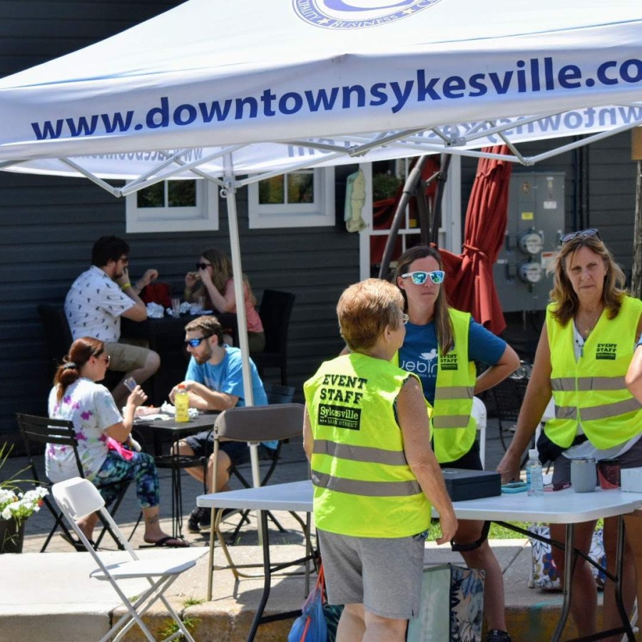 A group of four event staff wearing neon yellow safety vests stand under a white canopy tent labeled “www.downtownsykesville.com,” talking with a woman in a blue shirt. Behind them, a few people sit at an outdoor café table eating and drinking, and a dark gray building façade frames the scene.