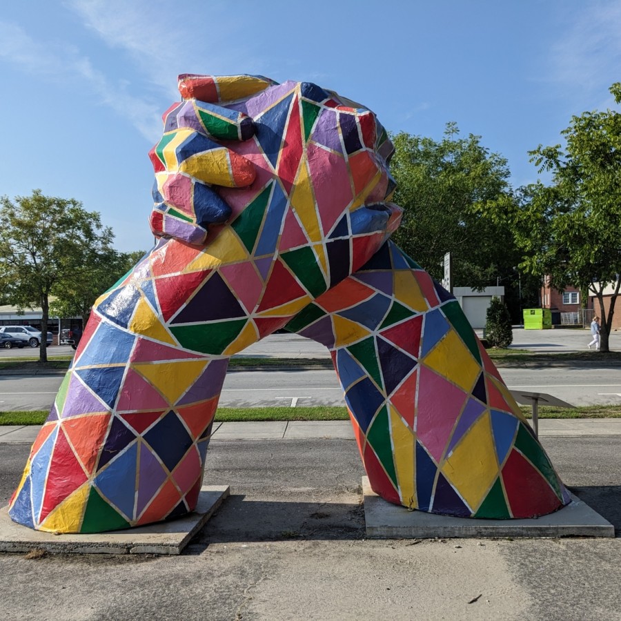 A large public sculpture of two oversized legs rising from square concrete bases and meeting at the top in a firm handshake. The entire piece is painted in a vibrant mosaic of geometric shapes—triangles and quadrilaterals in red, yellow, green, blue, pink, and purple—outlined in white. It stands on a roadside sidewalk with a parking lot, trees, and low buildings visible in the background.