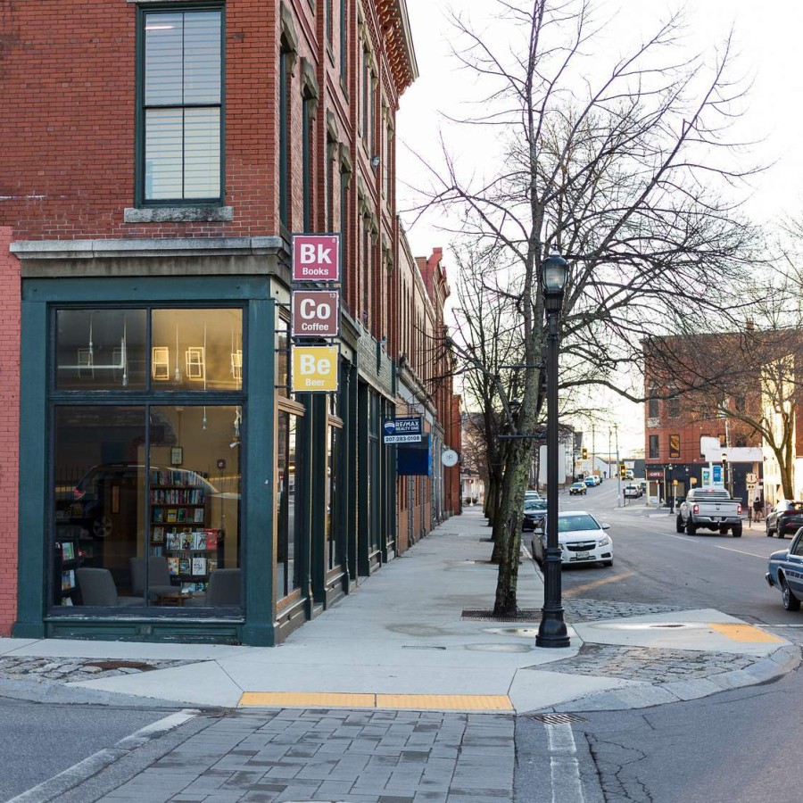 A quiet downtown corner at dusk shows a red-brick building with large display windows revealing bookshelves inside. To the left, a colorful geometric mural in shades of pink, yellow, and orange fills the lower brick wall. Vertical signs above the doorway read “Bk Books,” “Co Coffee,” and “Be Beer.” A leafless street tree and vintage streetlamp stand on the wide concrete sidewalk, while a blue-and-white classic sedan and other cars drift past on the two-lane road flanked by more historic storefronts.