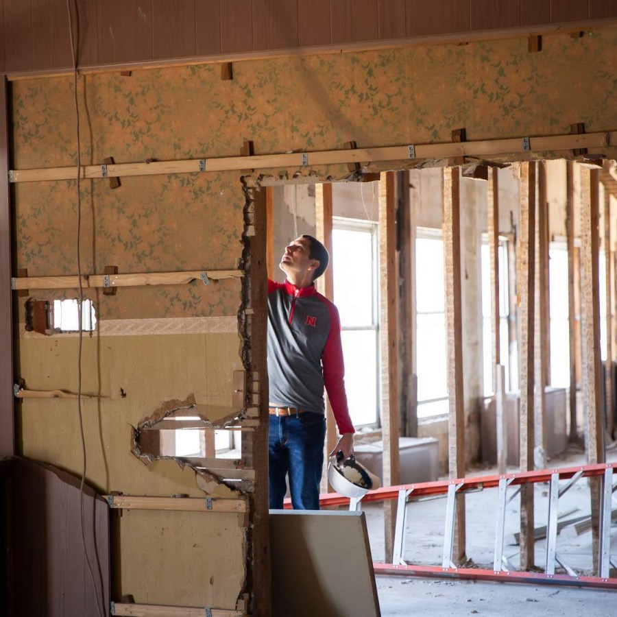 A person in a red-and-gray pullover stands inside a partially demolished interior, holding a white hard hat and looking up at exposed wooden studs and wall framing. The room has drywall removed from one side, a red ladder lying on the floor, and sunlight streaming through tall windows in the background.