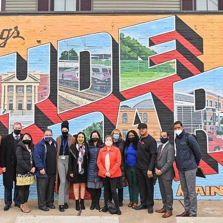 A group of about a dozen people in mixed business and casual attire stand side by side in front of a colorful brick-wall mural that reads “Greetings from Hyde Park,” with each letter filled by painted scenes of local landmarks. Most are wearing face masks, and a house’s siding is visible above the mural.