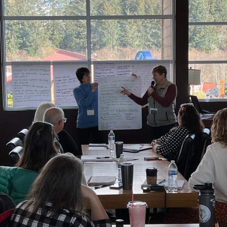 Two workshop facilitators stand at the front of a room holding a large flipchart covered in handwritten notes. One facilitator gestures toward the chart while speaking into a handheld microphone; the other holds the chart steady. In the foreground, participants sit at a long table with notebooks, water bottles, and coffee cups, watching and listening. Floor-to-ceiling windows behind the presenters look out onto a forested landscape.