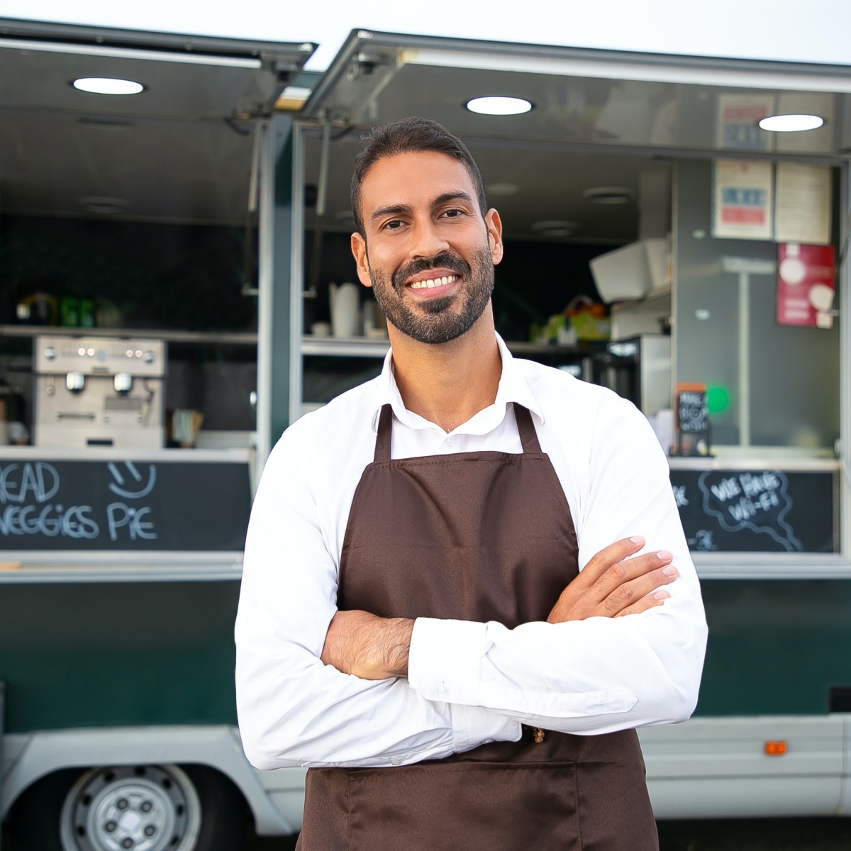 Un hombre con delantal marrón se cruza de brazos y sonríe frente a un camión de comida verde.