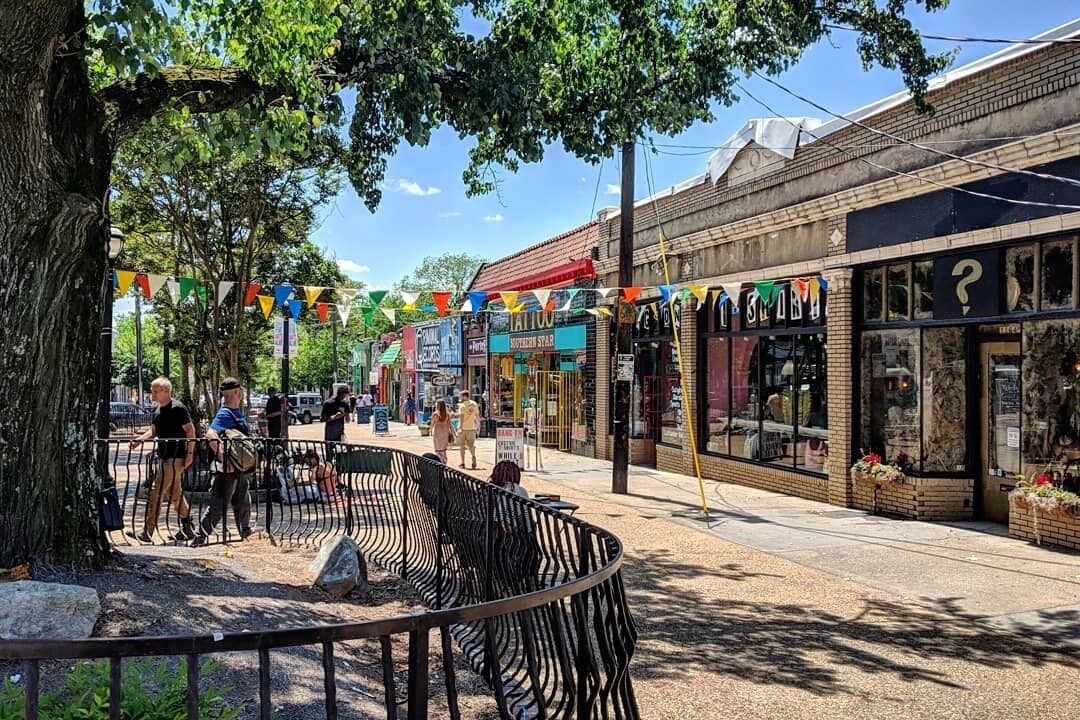 Little 5 Points Plaza Patio in Fulton County, Georgia, a commercial district with businesses and people walking on the sidewalk, with multi-colored flags hanging, trees lining the street.