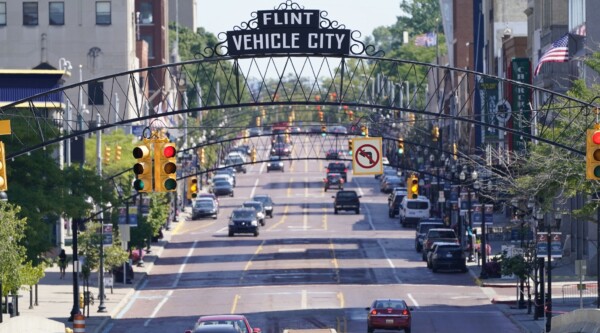 A street view of downtown Flint, Michigan, cars driving down the street, a sign reading, "Flint Vehicle City" is over the road.