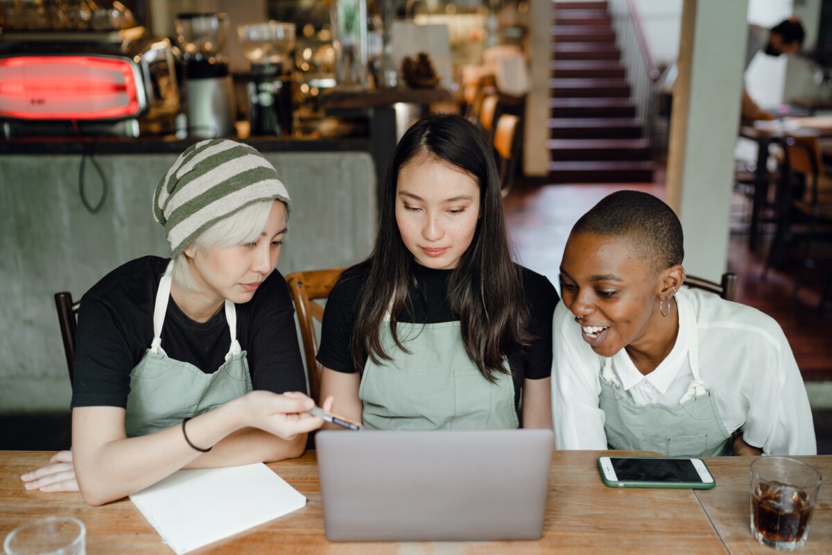 A group of three women in aprons gather around a computer in a coffee shop.