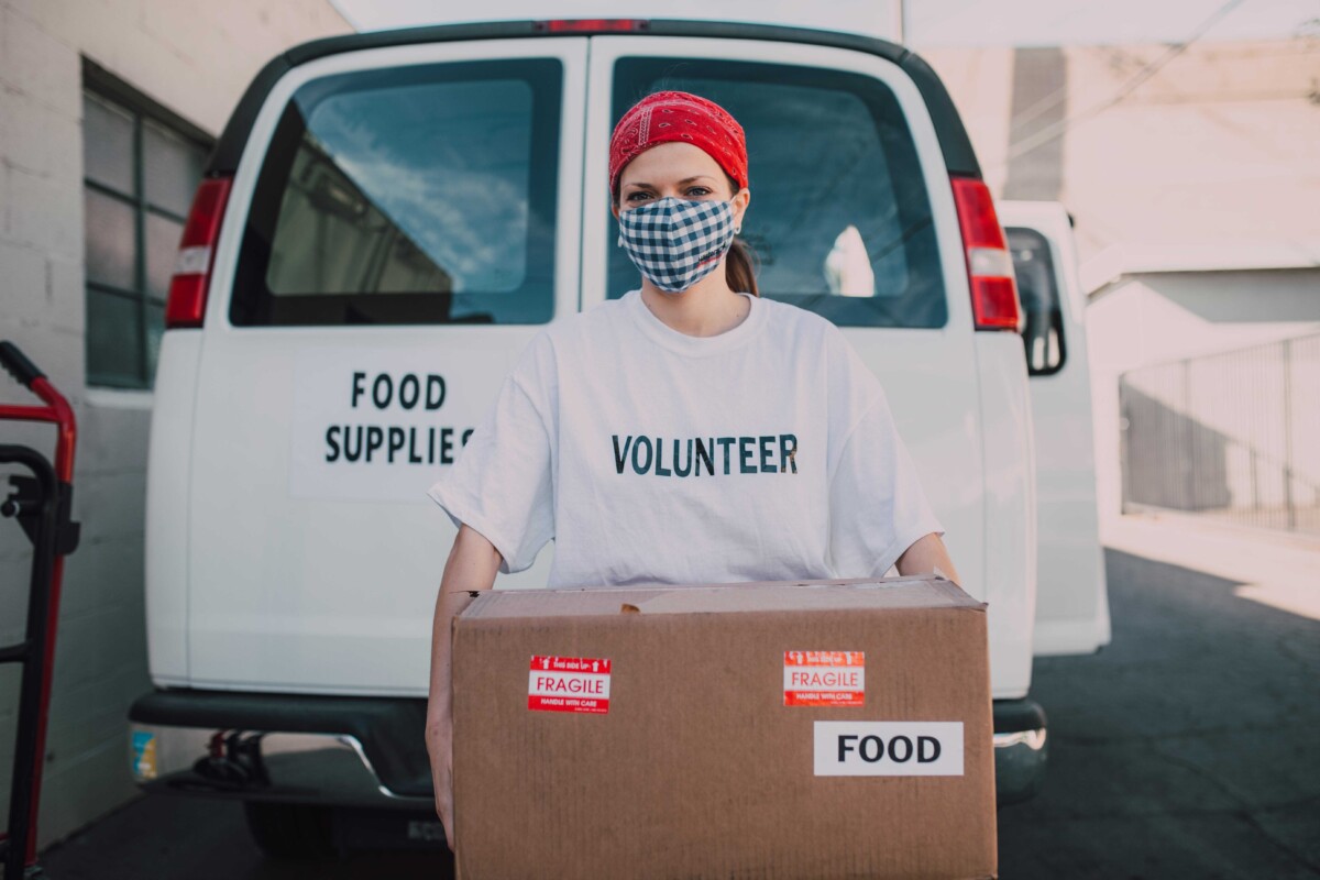 Una voluntaria con una cinta roja en la cabeza y una mascarilla sostiene una caja de cartón con la etiqueta "alimentos" delante de una furgoneta blanca con la etiqueta "suministros alimentarios".