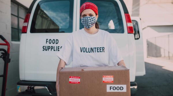 Una voluntaria con una cinta roja en la cabeza y una mascarilla sostiene una caja de cartón con la etiqueta "alimentos" delante de una furgoneta blanca con la etiqueta "suministros alimentarios".