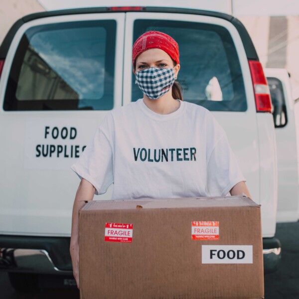 A woman volunteer with a red headband and face mask holds a cardboard box labeled "food" in front of a white van labeled "food supplies."