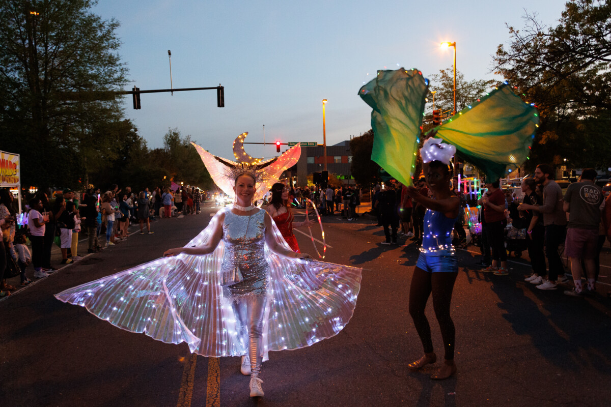 During a parade at dusk, a woman wearing a light-up dress and wings walks down a city street.