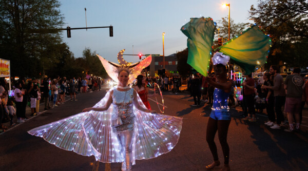 Durante un desfile al atardecer, una mujer con un vestido iluminado y alas camina por una calle de la ciudad.