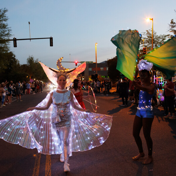 During a parade at dusk, a woman wearing a light-up dress and wings walks down a city street.