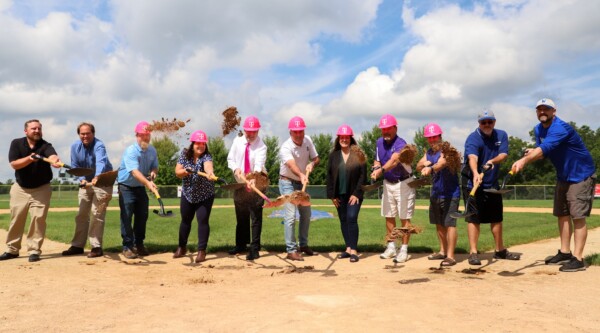 A group of people wearing T-Mobile branded hard hats and holding shovels gather on a baseball field.