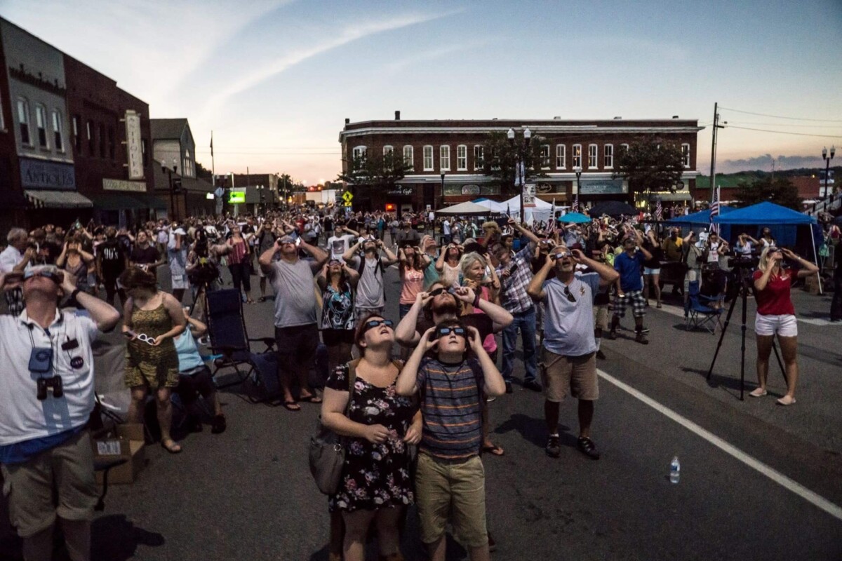 Photo of a crowd of people wearing eclipse protective eyeglasses look up at the sky in a downtown scene.