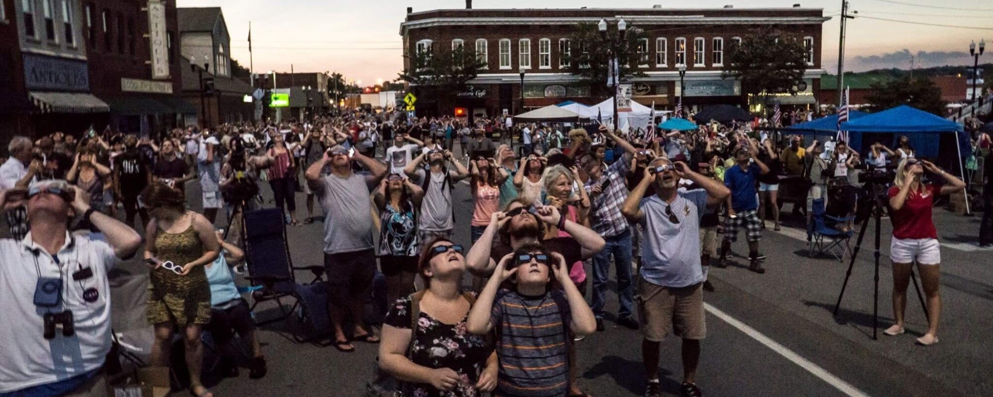 Photo of a crowd of people wearing eclipse protective eyeglasses look up at the sky in a downtown scene.