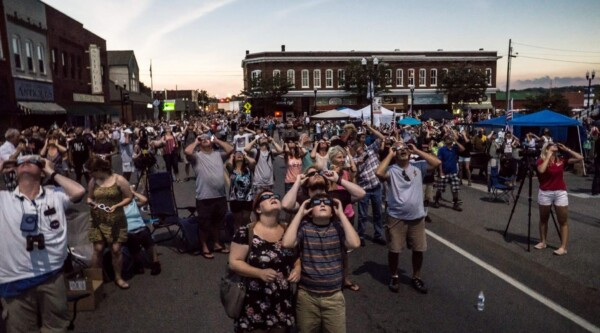 Photo of a crowd of people wearing eclipse protective eyeglasses look up at the sky in a downtown scene.