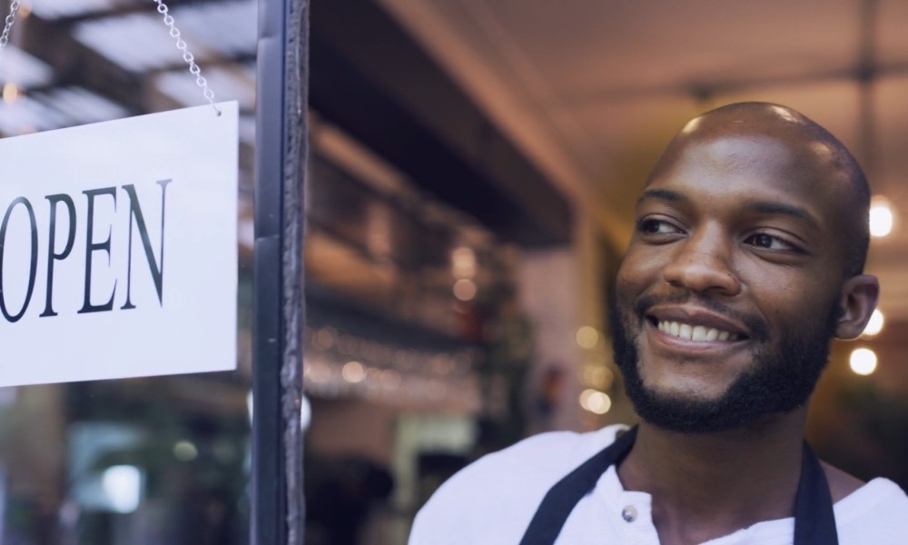 Where It Starts: Breaking Barriers to Business, an initiative of the Truist Foundation, video thumbnail. A smiling business owner opens the door to his business. An "Open" sign hangs on the door.