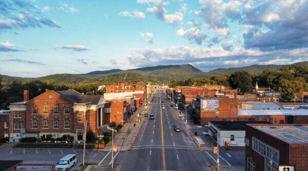 A photo of a downtown with brick buildings, four lane street, a hilly landscape, and a blue sky with clouds.