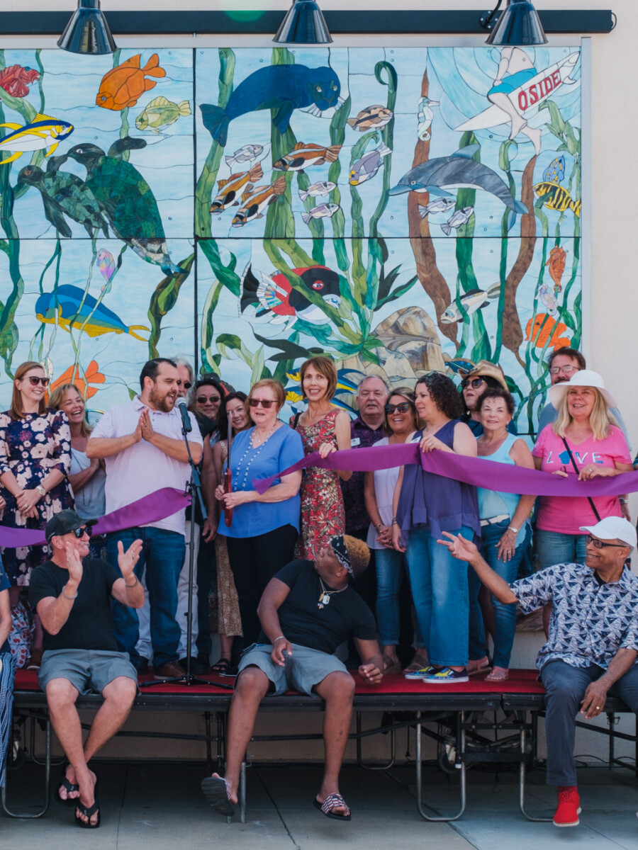 A large group of adults and children gather on a stage for a ribbon cutting event celebrating the completion of a nautical-themed mosaic mural that is visible behind them.