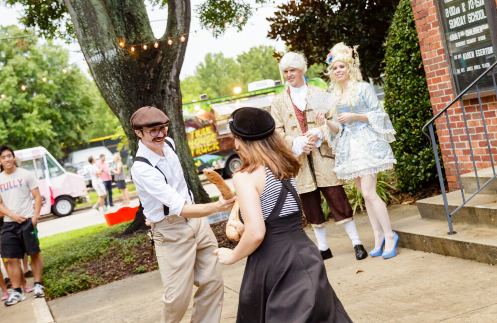 Two people spar with baguettes while a couple dressed in 18th century French court-inspired attire.