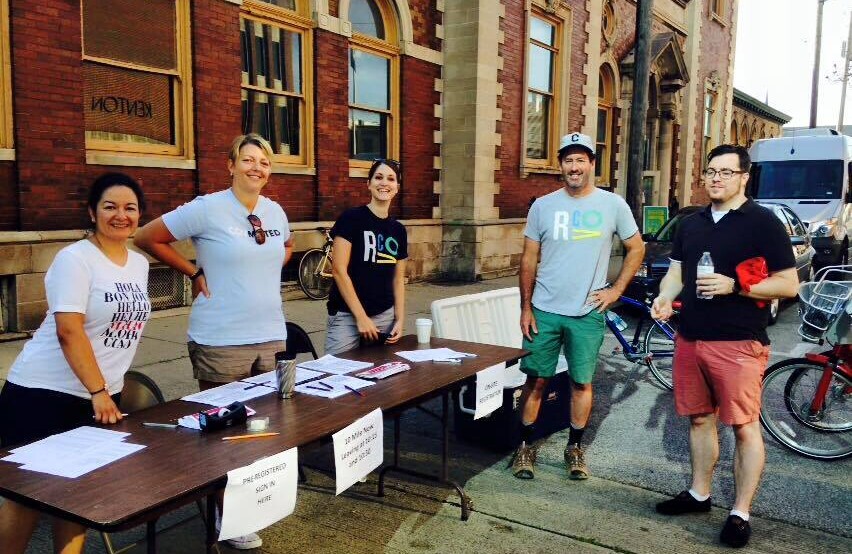 Five people stand around a registration table set-up in front of a brick building.