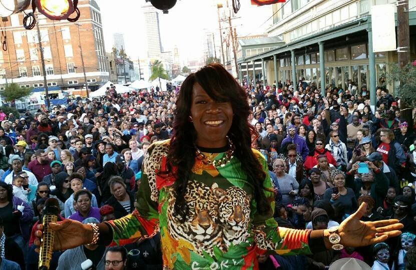 A woman smiles while holding a microphone with her back turned towards a large crowd that has gathered in front of the stage she is standing on.