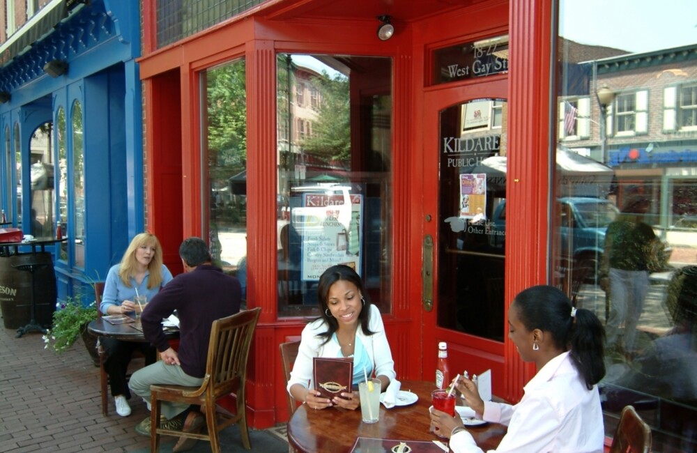 People sit at sidewalk dining tables outside an eatery.