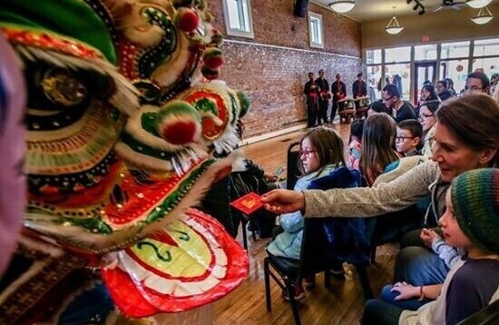 Children watch as a woman extends a Chinese New Year red envelope towards the open mouth of a dancing lion.