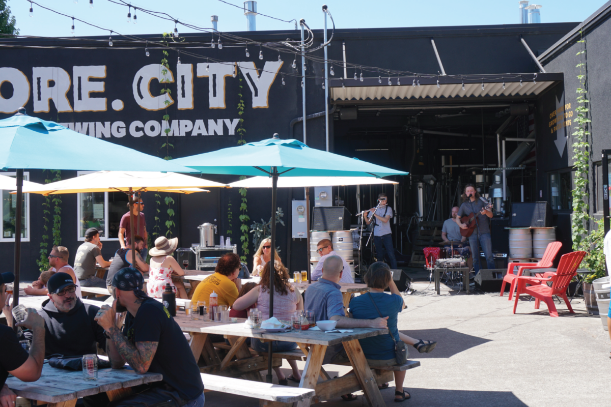 On a sunny day people gather at a brewery's outdoor picnic tables.