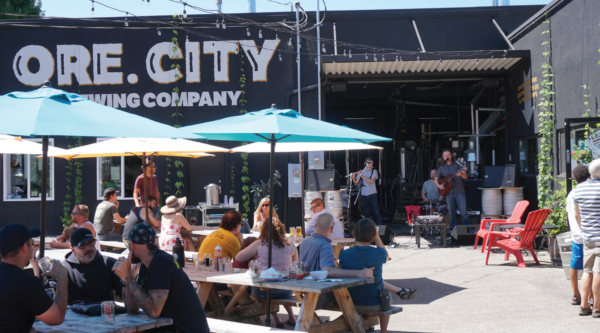 On a sunny day people gather at a brewery's outdoor picnic tables.