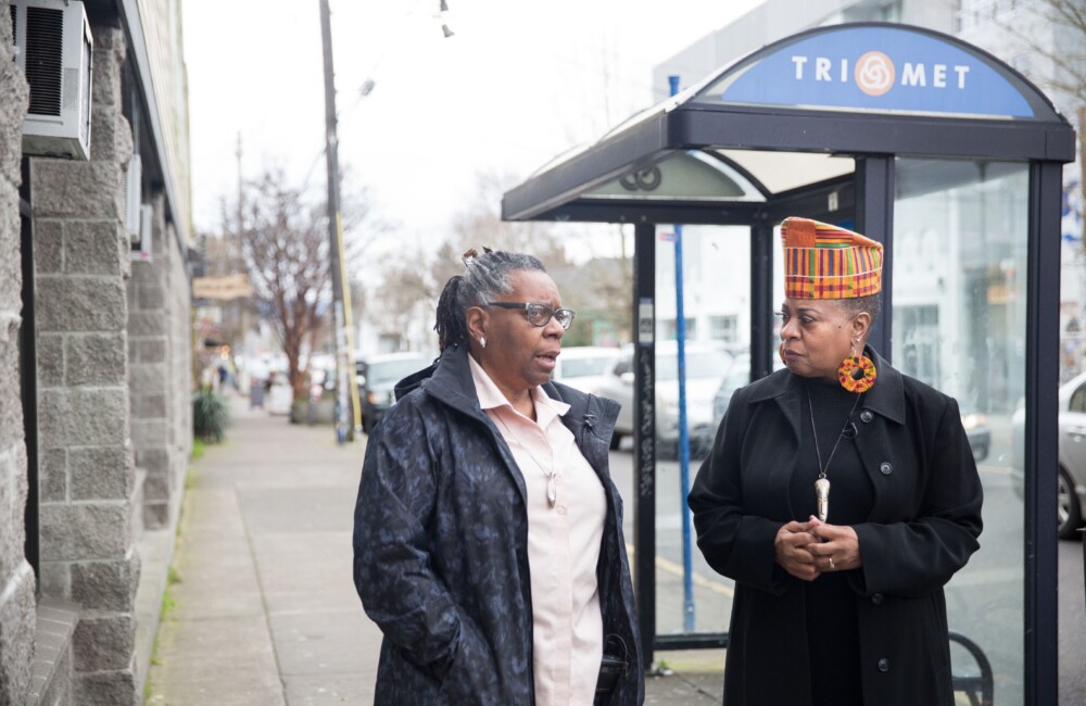 Two women engage in conversation while standing near a bus stop.