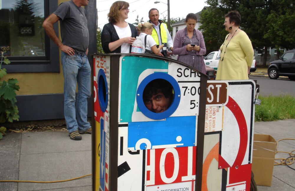 A man playfully peers from inside a trash receptacle made from repurposed street signs.